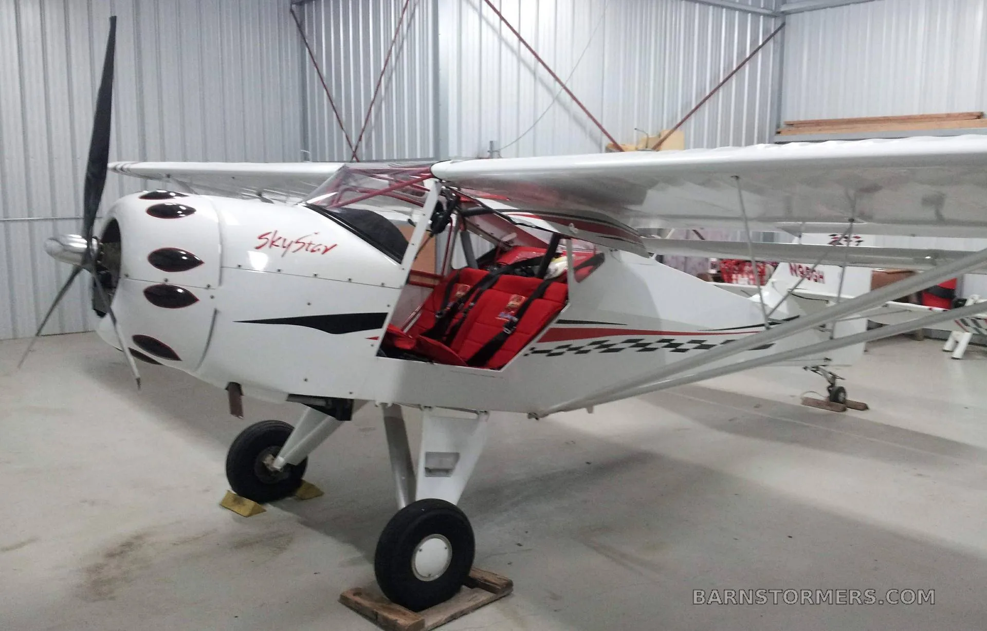 A white SkyStar Kitfox aircraft with red interior and open door is parked inside a hangar.