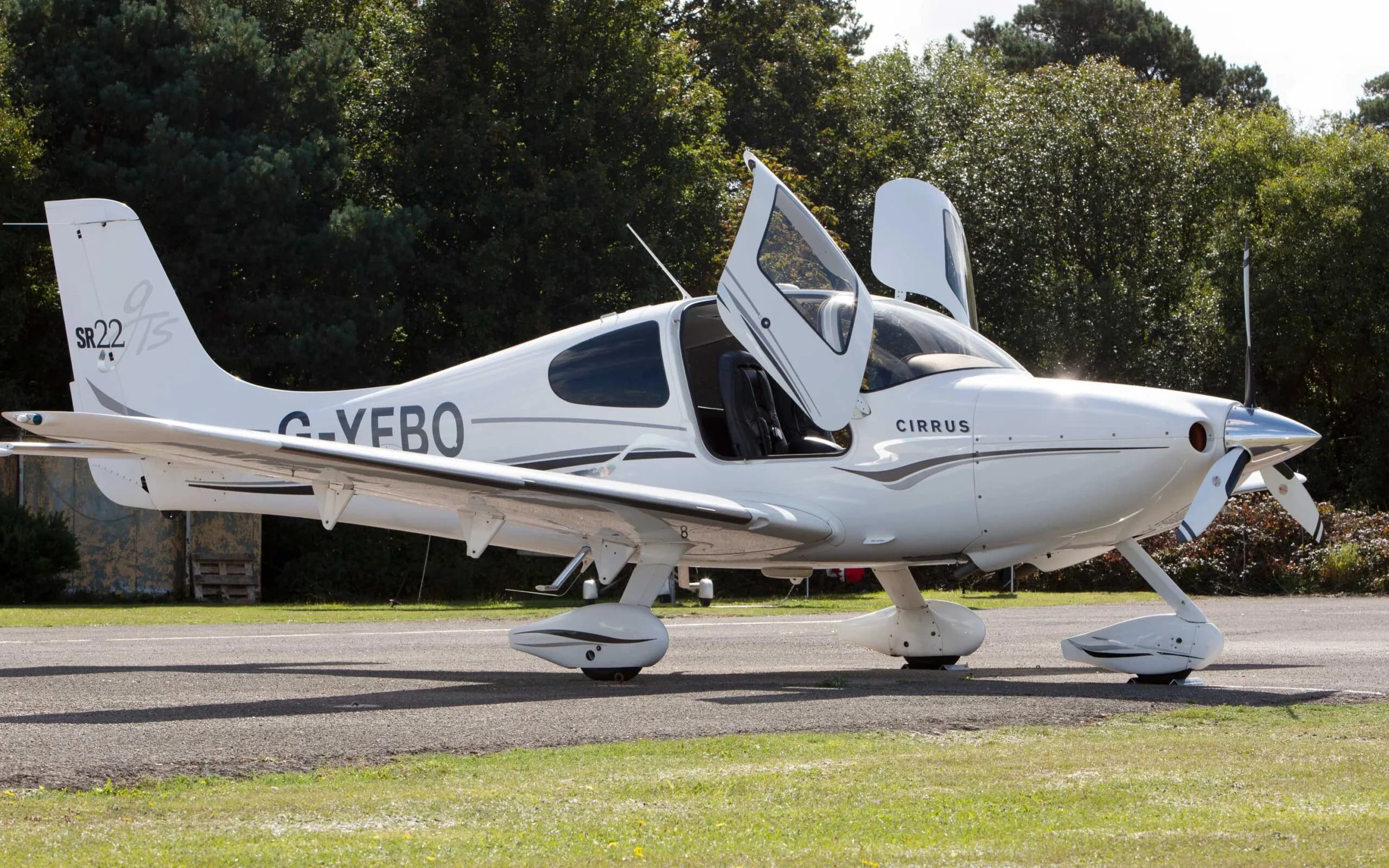 A white Cirrus SR22 TSi aircraft with black and grey stripes, registration G-YEBO, parked on an airfield with its gull-wing doors open.