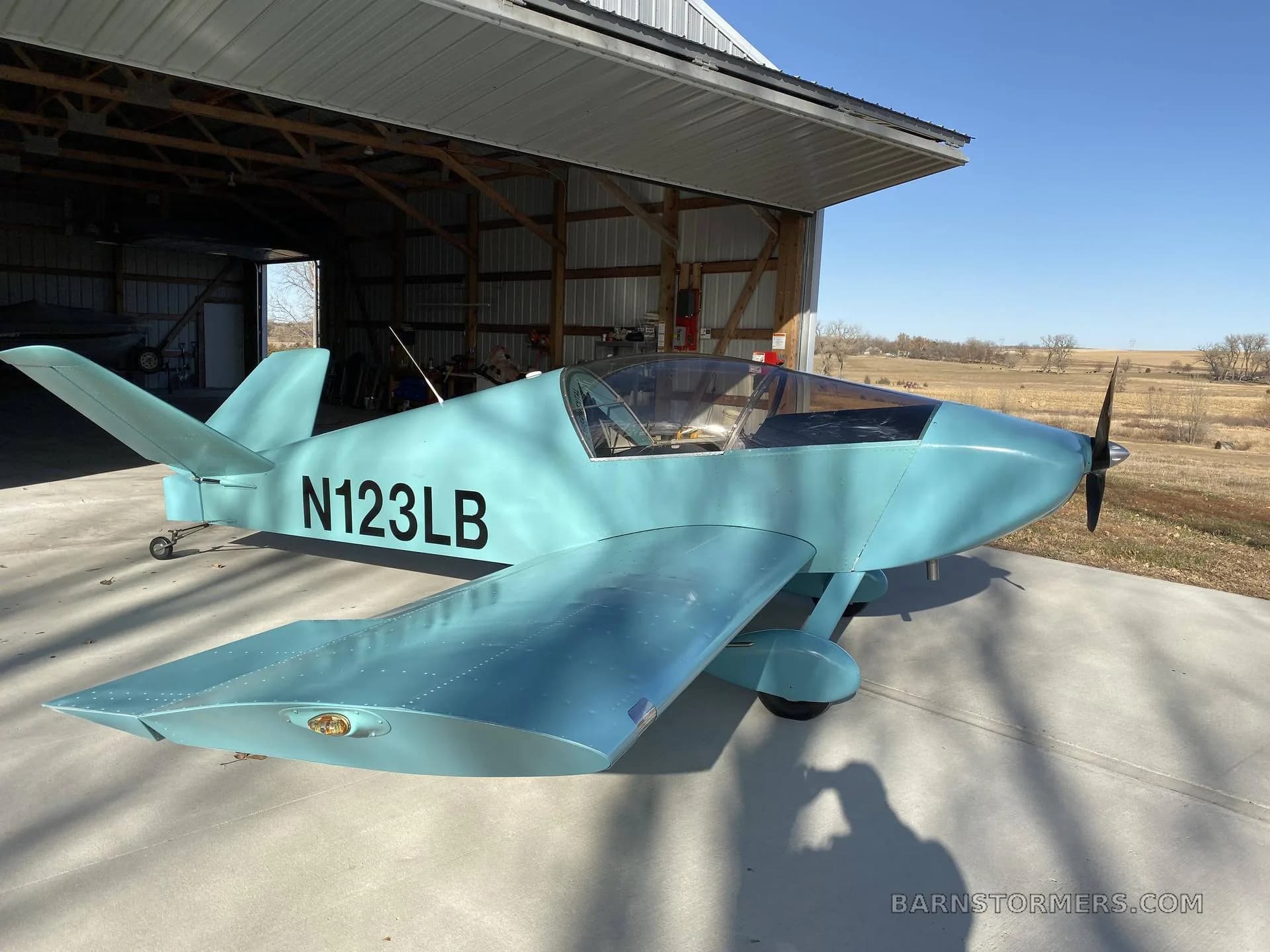 A light blue single-engine experimental aircraft (N123LB) with a bubble canopy, parked outside a hangar on a sunny day.