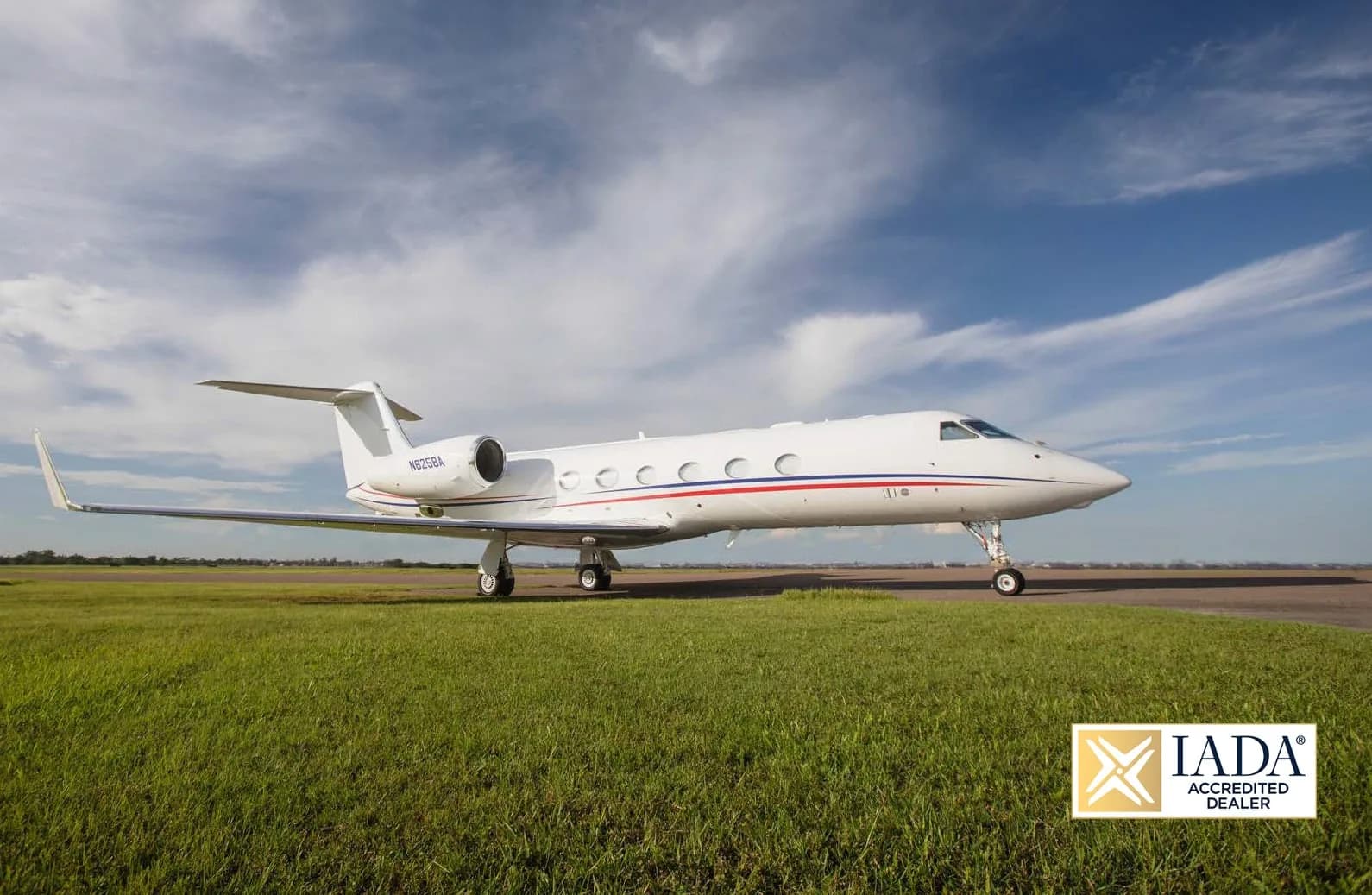 A white Gulfstream G650 business jet with red and blue stripes on the fuselage, parked on a runway with grass.