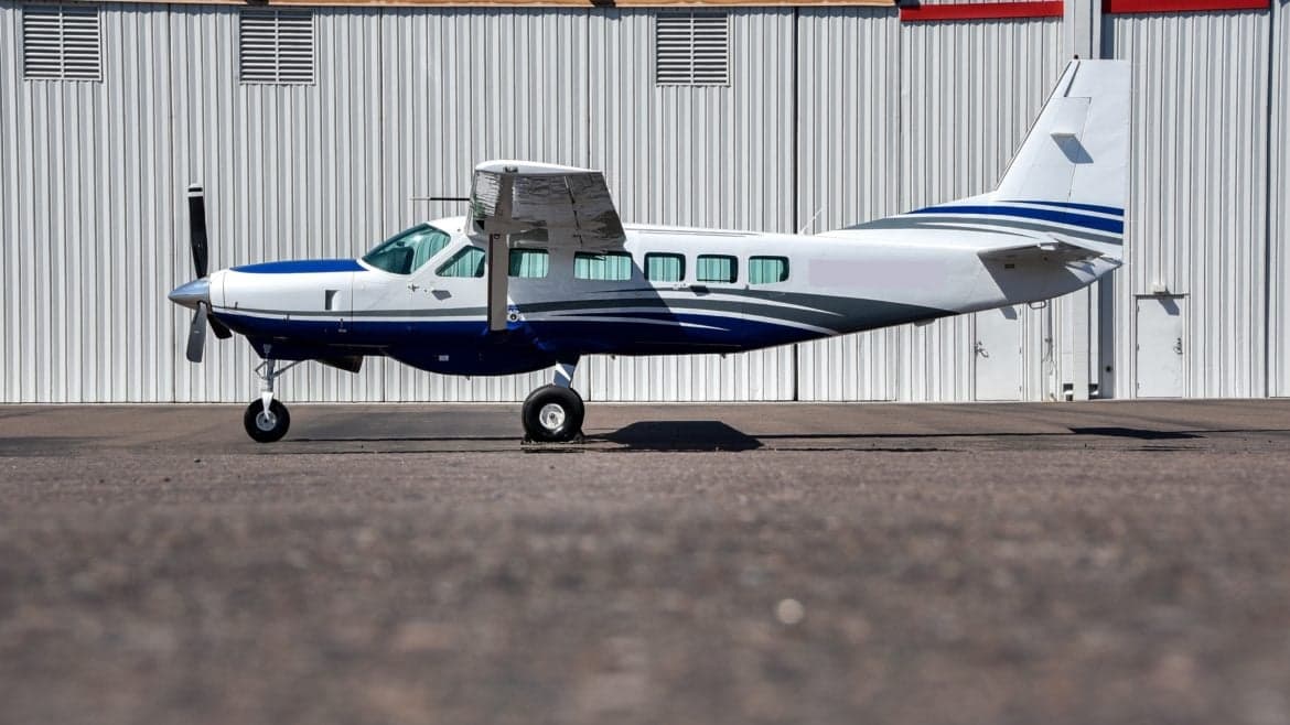 A side view of a blue and white Cessna Caravan on the tarmac in front of a hangar.