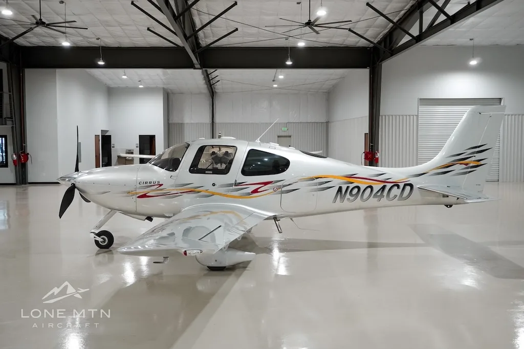 A silver Cirrus aircraft with red and yellow accent stripes, registration N904CD, parked in a hangar.