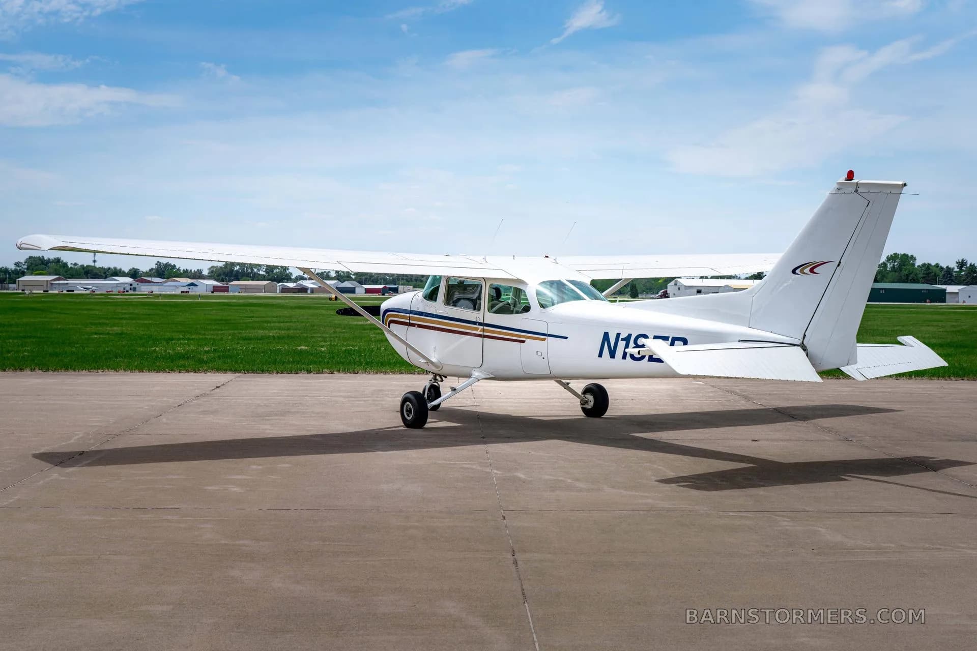 A white Cessna 172 (N18EB) with blue and gold stripes is parked on a tarmac, viewed from the rear-right at a distance.