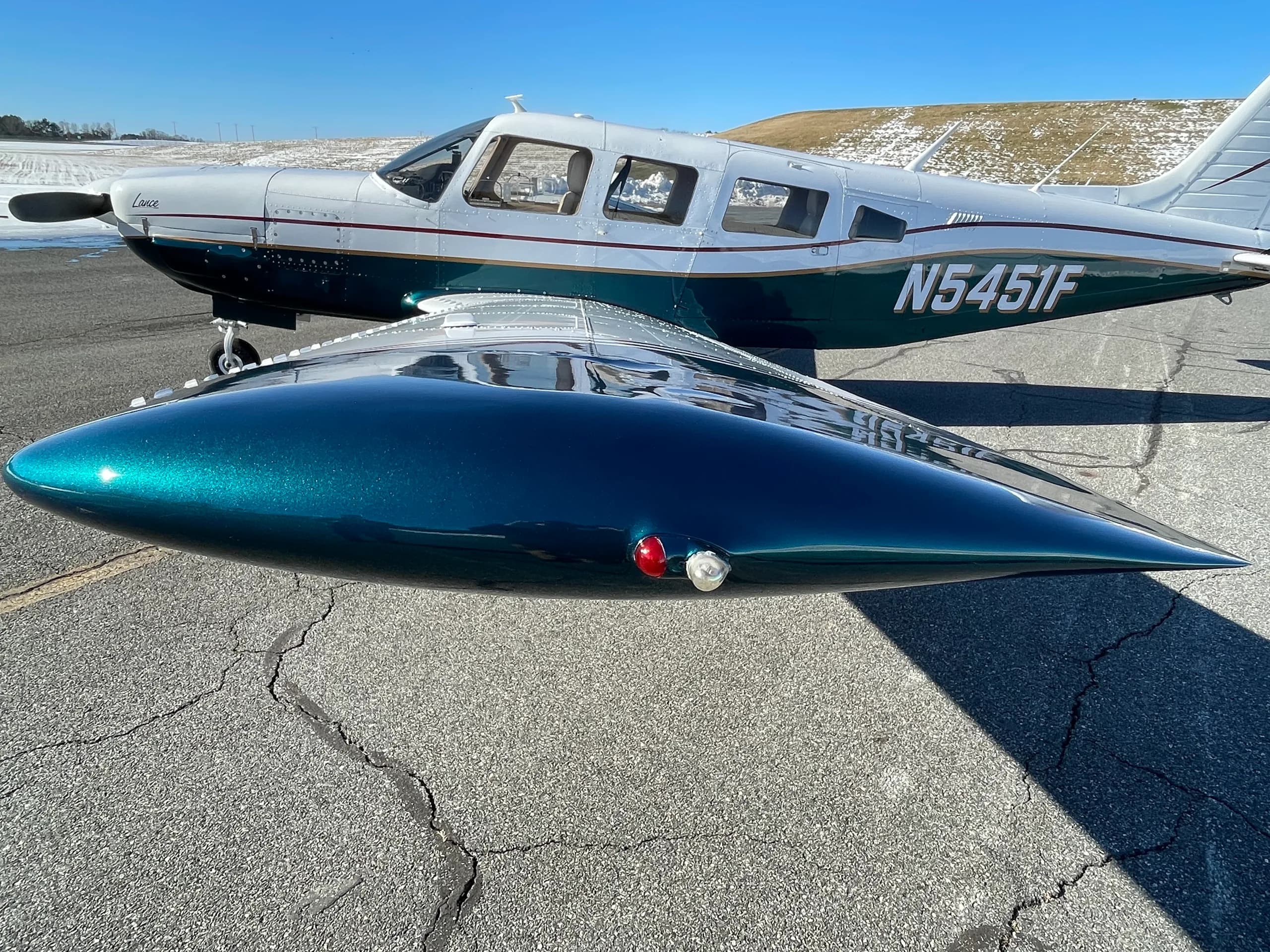 A white and dark green Piper Lance aircraft, registration N5451F, parked on an asphalt ramp with a snowy field in the background.
