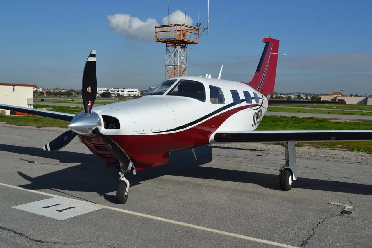A white and red single-engine propeller aircraft, possibly a Piper Malibu, is parked on an airfield ramp with a tower in the background.