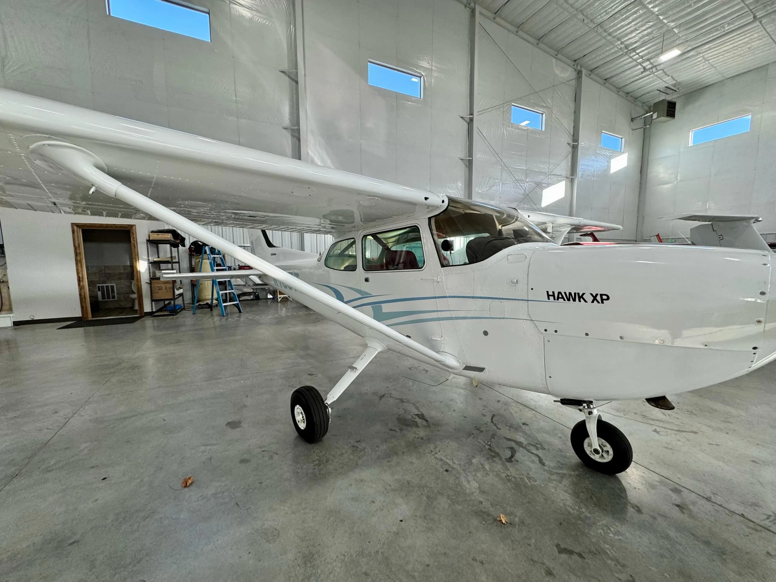 A three-quarter front view of a white Cessna 172 Hawk XP aircraft with blue stripes, parked in a hangar.