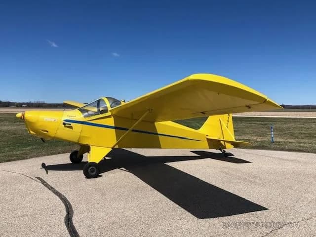 A bright yellow single-engine experimental aircraft with blue stripes, viewed from the left side, parked on an asphalt surface.