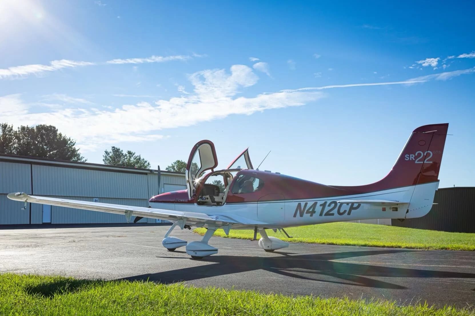 A red and white Cirrus SR22 GTS aircraft (N412CP) with its doors open, parked on an airfield under a sunny sky.