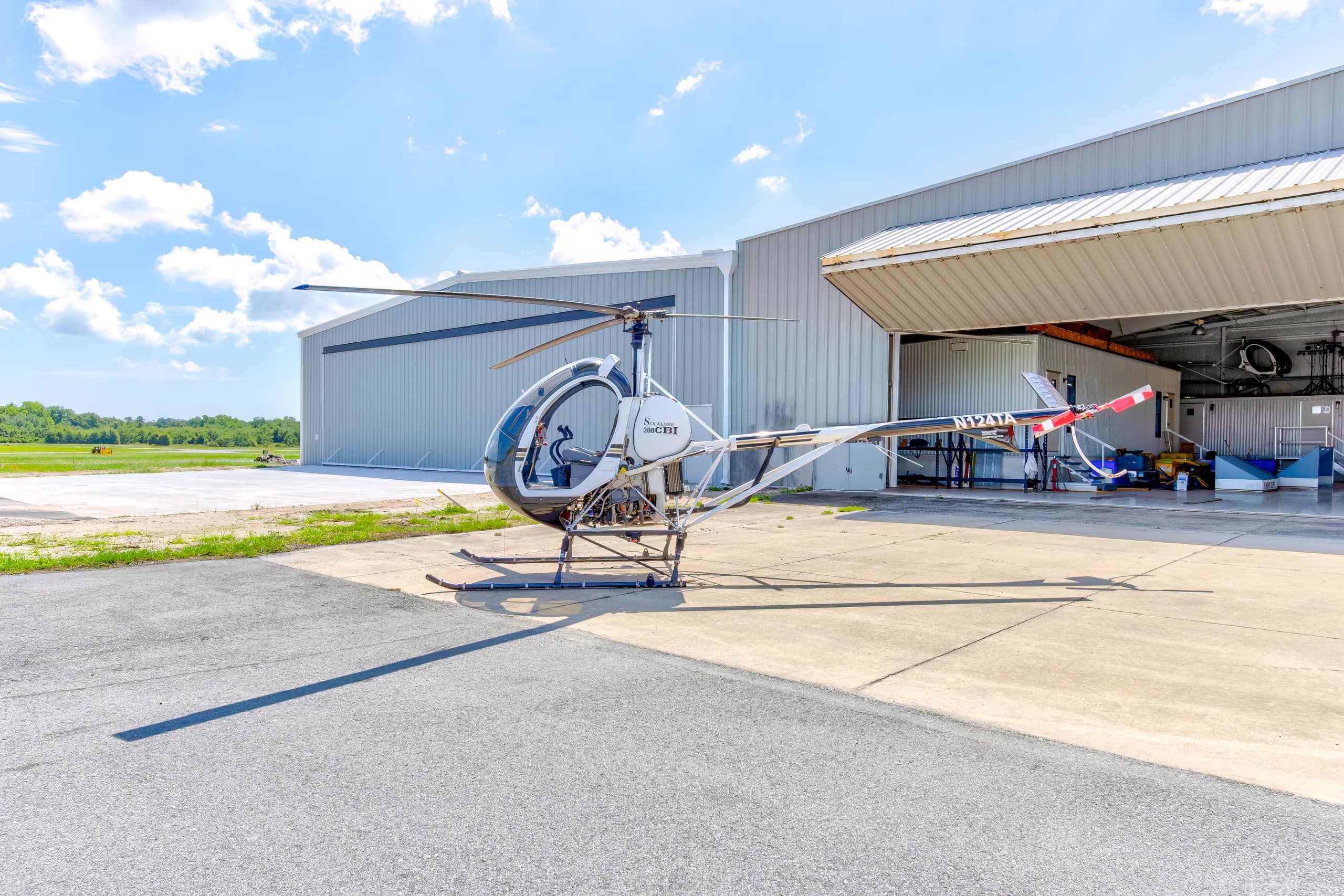A white and black Schweizer 300CBi helicopter, N124TA, parked on an airfield tarmac next to a hangar under a blue sky.