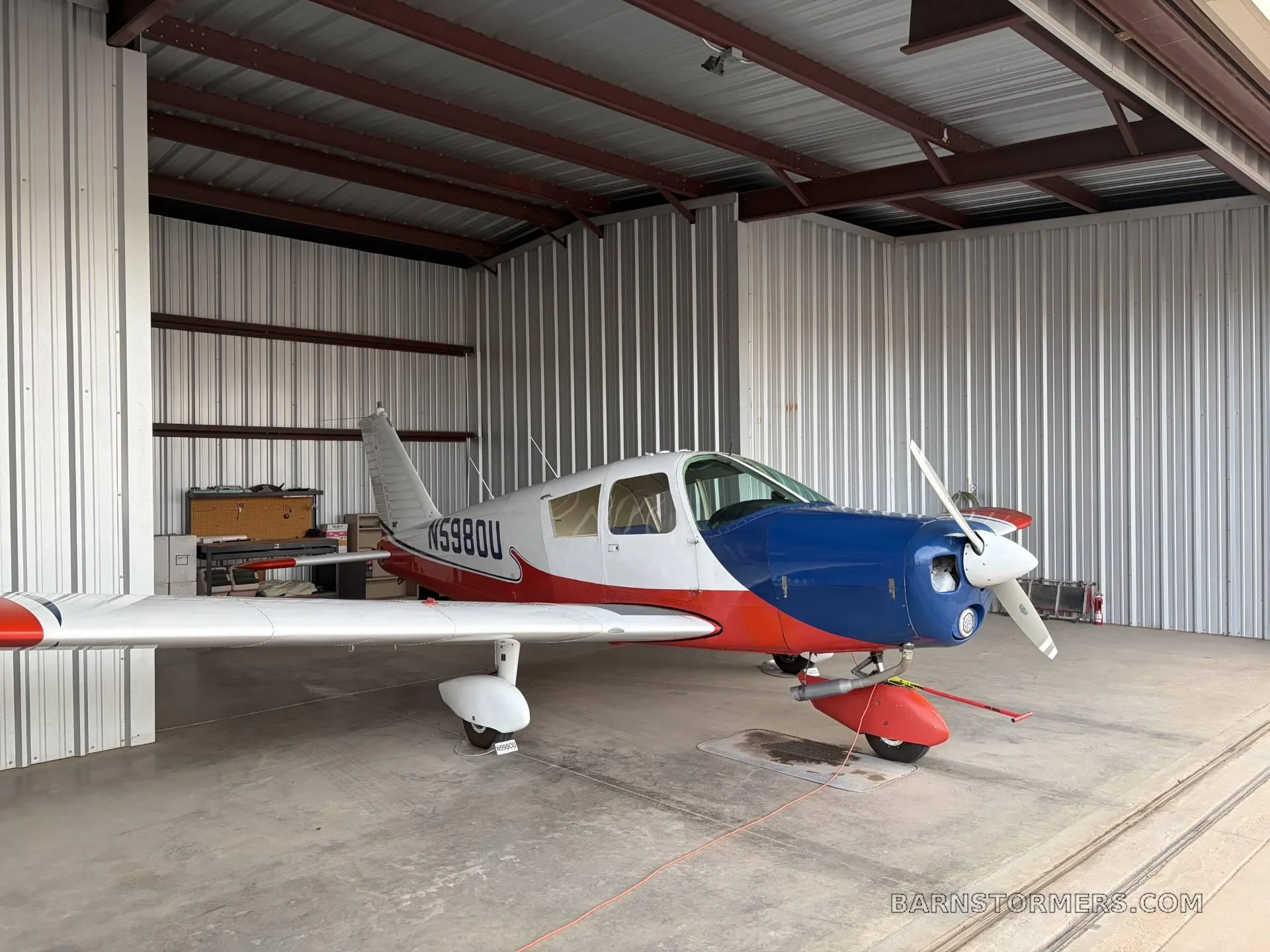 A red, white, and blue single-engine propeller aircraft (N5980U) is parked inside a metal hangar.