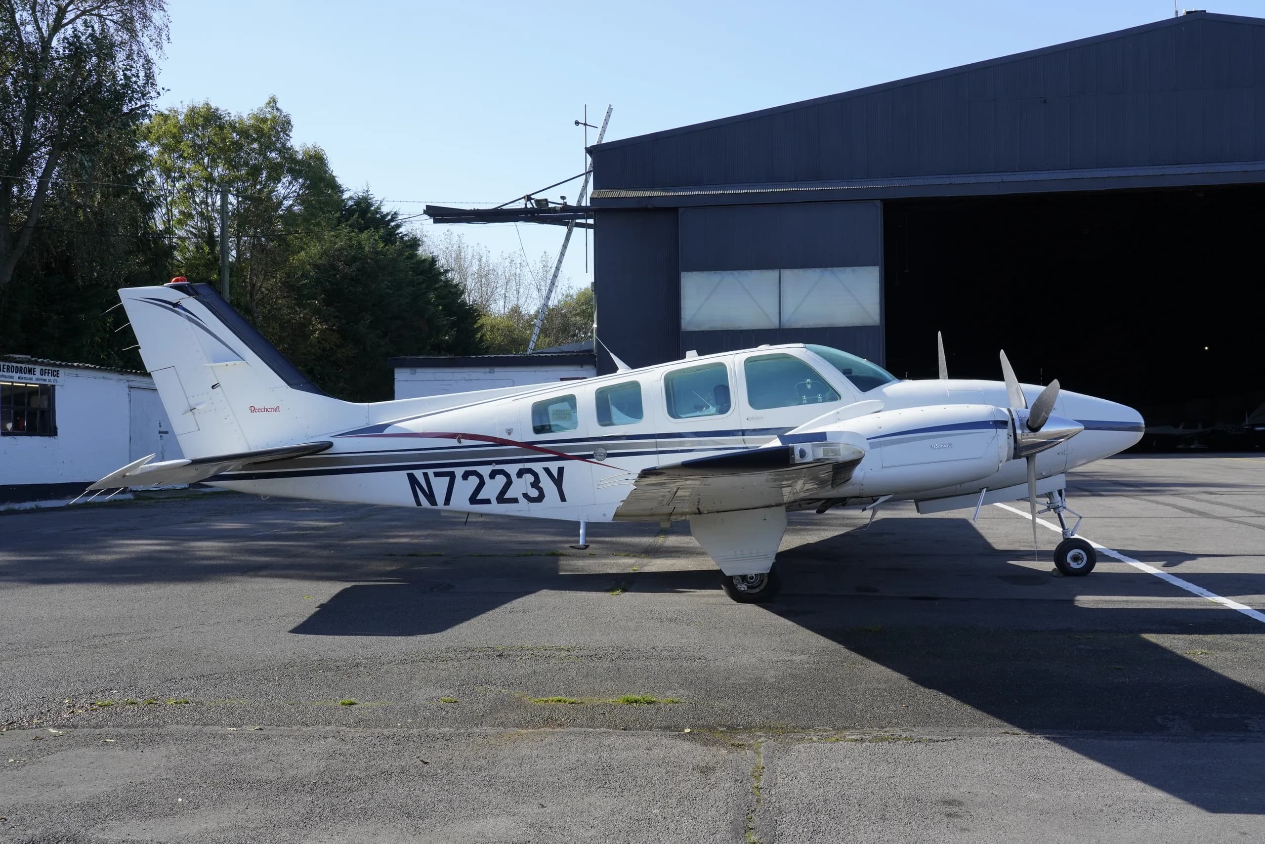 A white twin-engine propeller aircraft with blue and black stripes and registration N7223Y parked on tarmac next to a large hangar.
