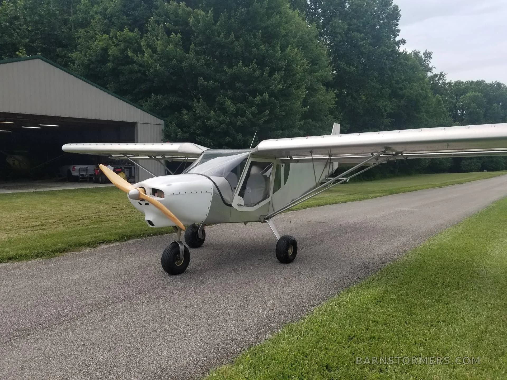 A light gray ultralight aircraft with a wooden propeller is parked on a paved path, with an open hangar and trees in the background.