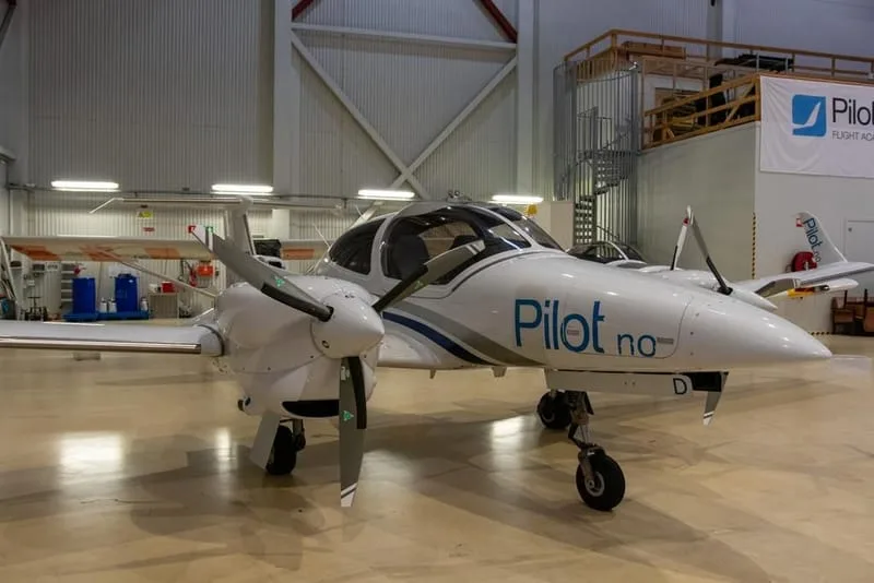 A white Diamond DA42 twin-engine aircraft with "Pilot.no" livery parked in a hangar.