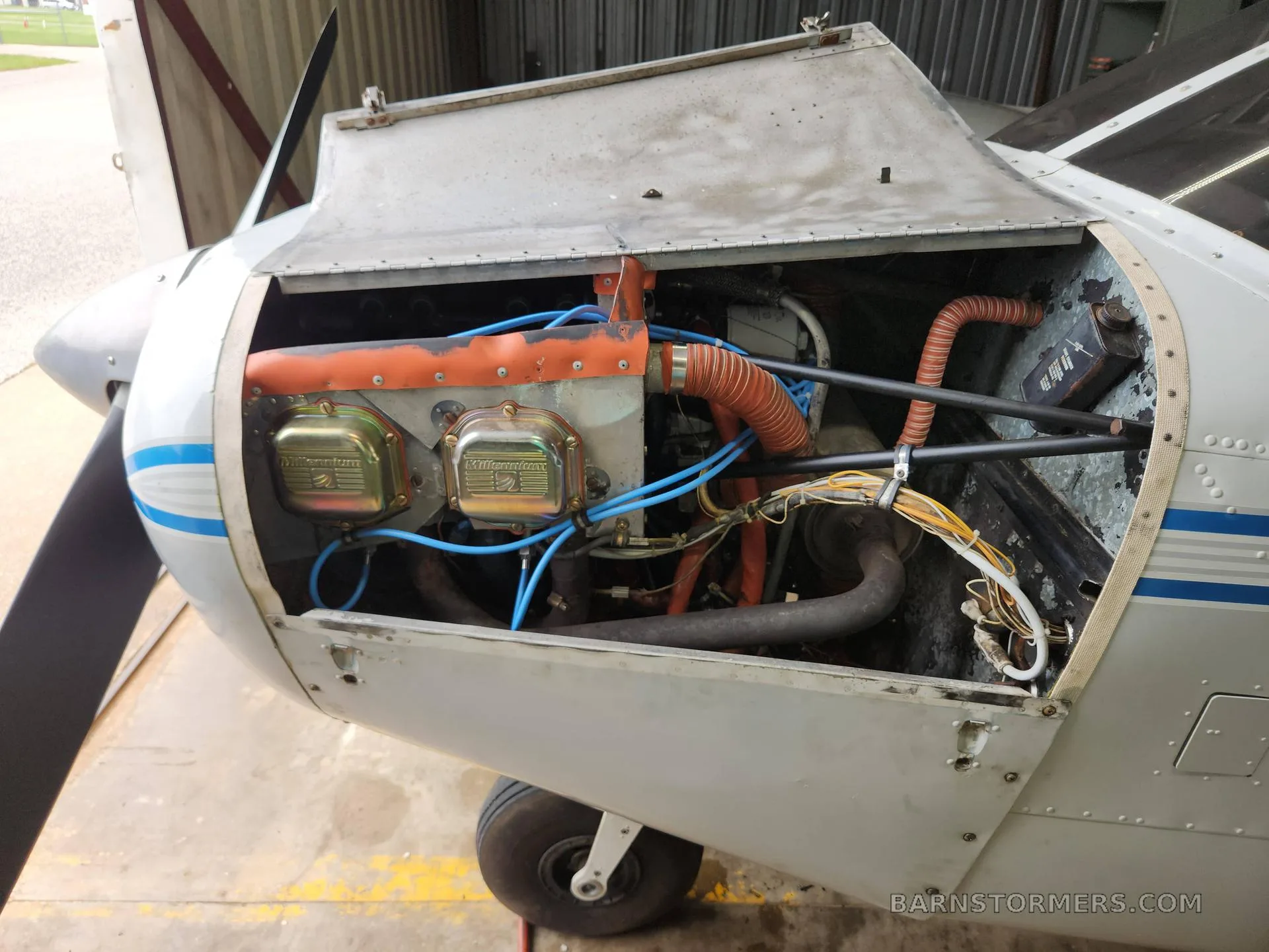 The exposed engine compartment of a Piper Cherokee aircraft, showing the engine with blue and orange hoses.