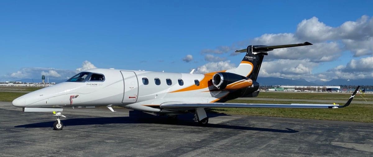 A Phenom 300 aircraft is parked on the tarmac with a white, grey, and orange paint scheme.