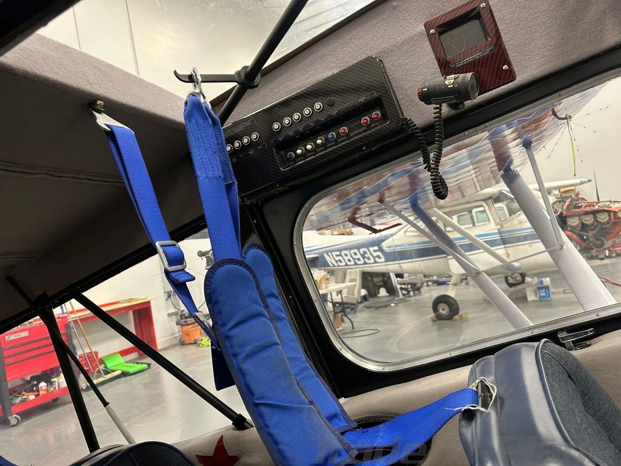 The interior of an aircraft cabin, showing a blue seat with harness and an overhead panel.