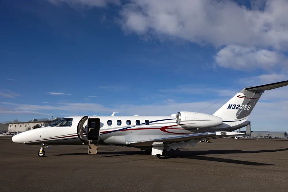 A white Cessna Citation business jet with red and blue stripes, registration N322SB, parked on an tarmac with its main door open.