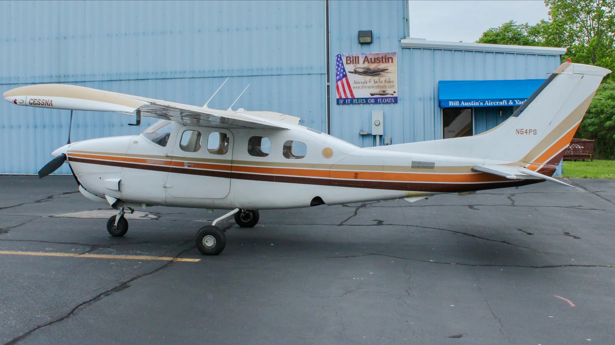 A side view of the white Cessna P210N Centurion, registration N54PS, with brown and orange stripes, parked next to a blue hangar.