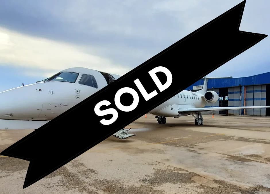 A white business jet is parked on an airport apron with a blue hangar in the background under a cloudy sky.