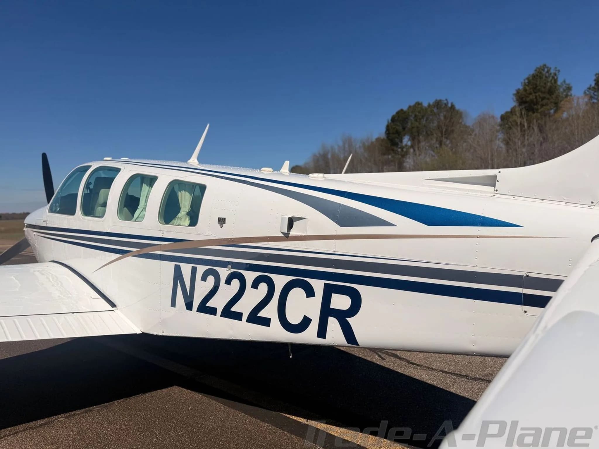 A close-up side view of the white Beechcraft Bonanza N222CR fuselage, highlighting the blue and gold paint scheme.