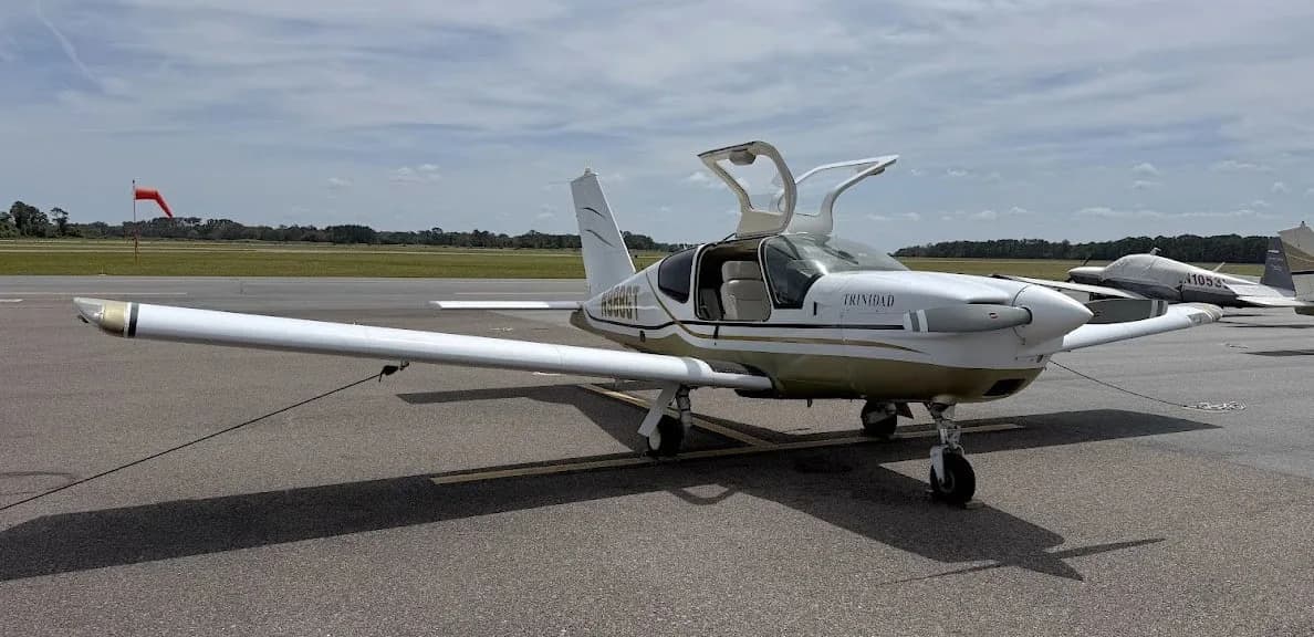 A white and gold Socata TB-20 Trinidad aircraft with its doors open is parked on an airfield next to another aircraft.