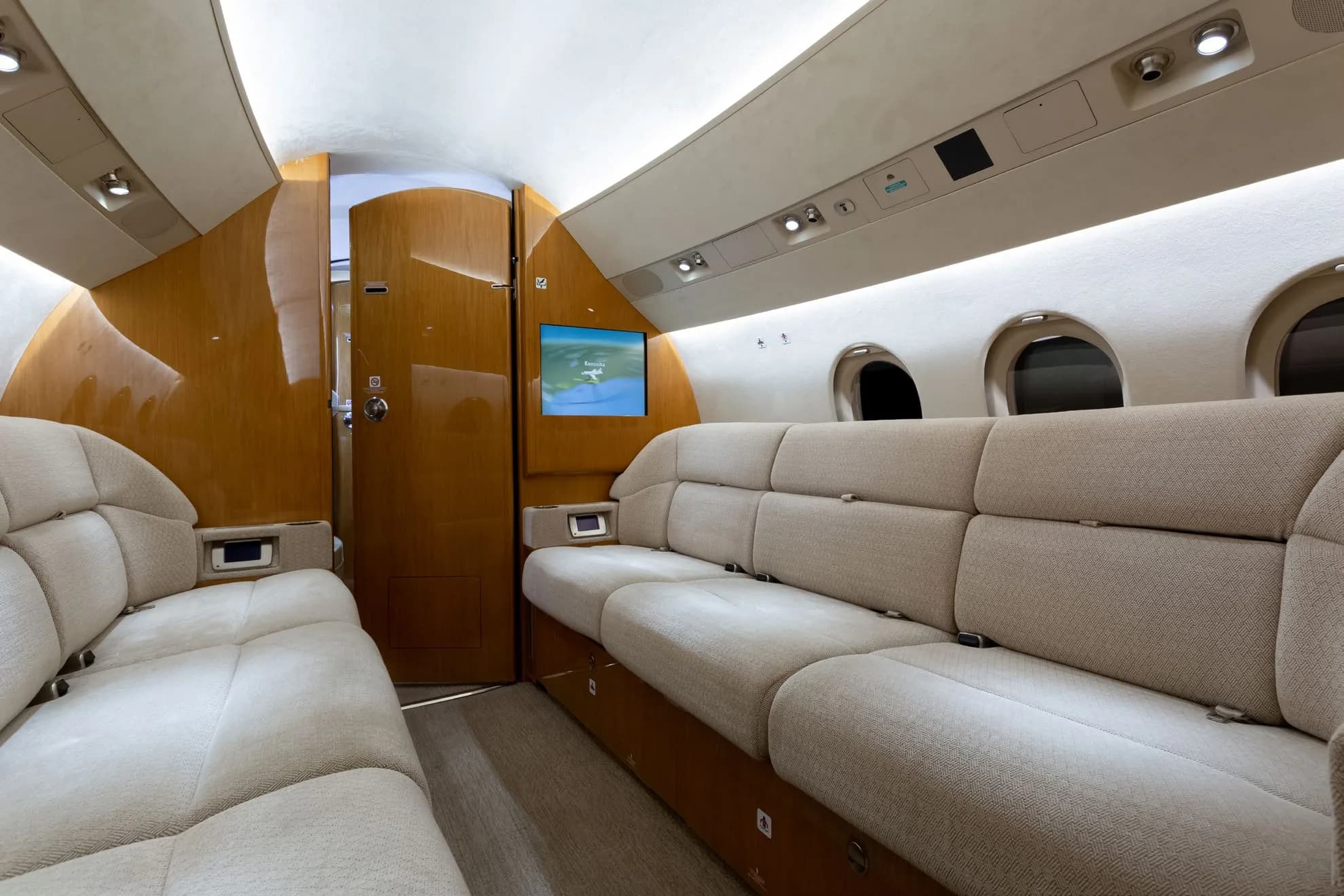 The spacious interior of a private jet cabin featuring two long beige fabric sofas and a screen displaying a map.