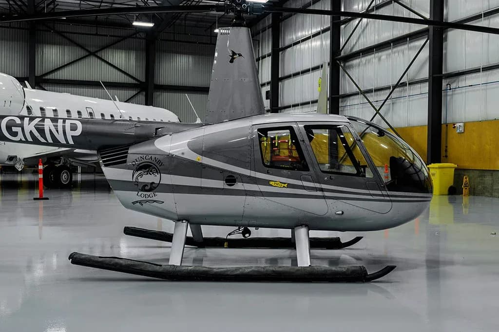 A side view of a grey and white Robinson R44 helicopter with "DUNCANBY LODGE" logo, parked in a hangar.