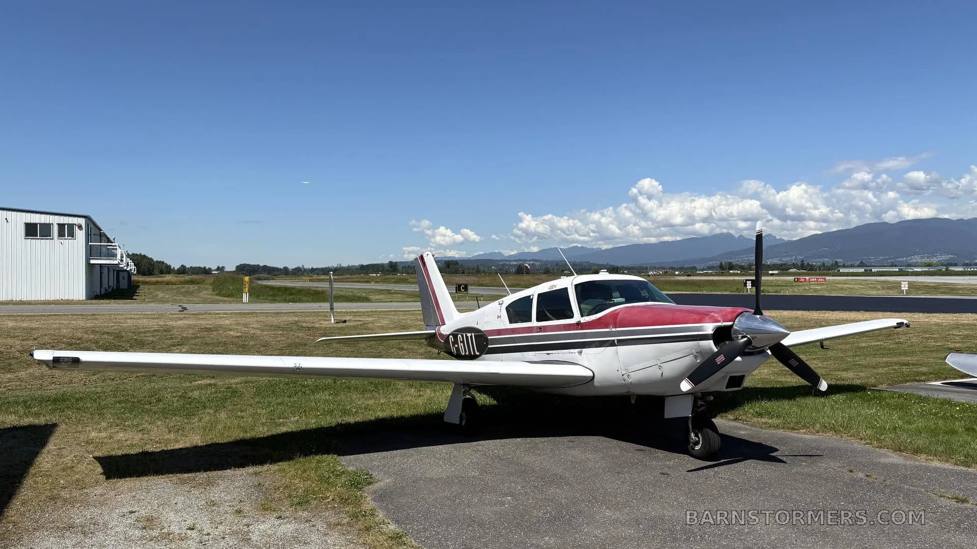 A white and red single-engine propeller aircraft (C-GJTL) parked on an asphalt ramp at an airfield with mountains in the background.