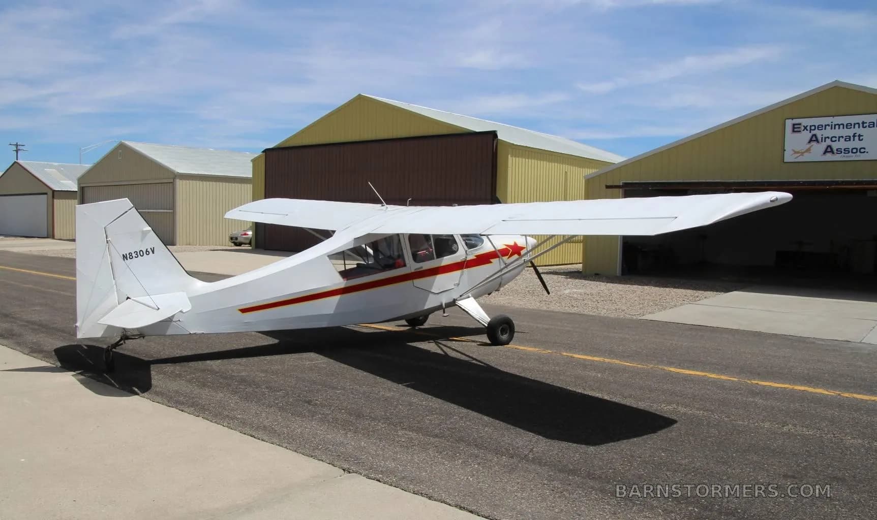 A white single-engine high-wing aircraft with red stripes and a red star on the cowl, seen from the rear-left, parked on an asphalt taxiway near hangars.