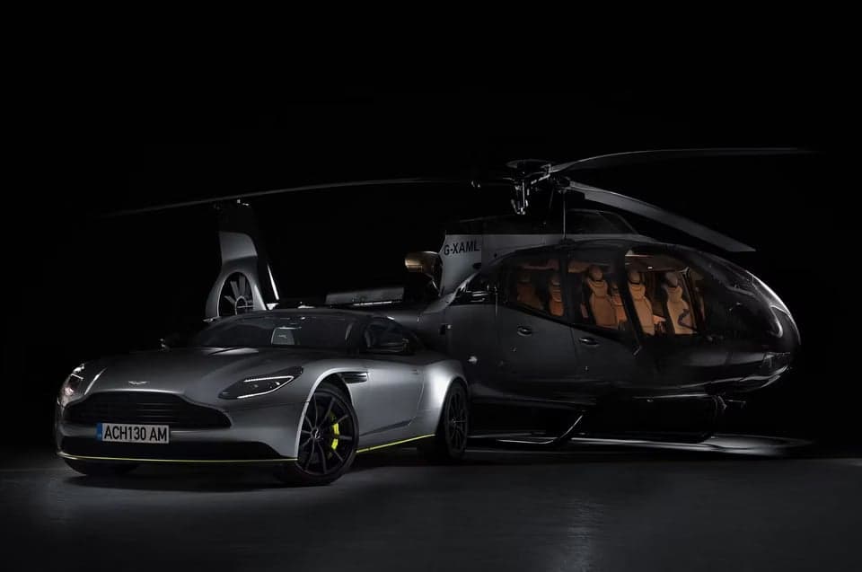 A dark helicopter parked next to a silver Aston Martin car.