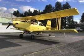 A yellow biplane crop duster sits on the tarmac in front of a building.