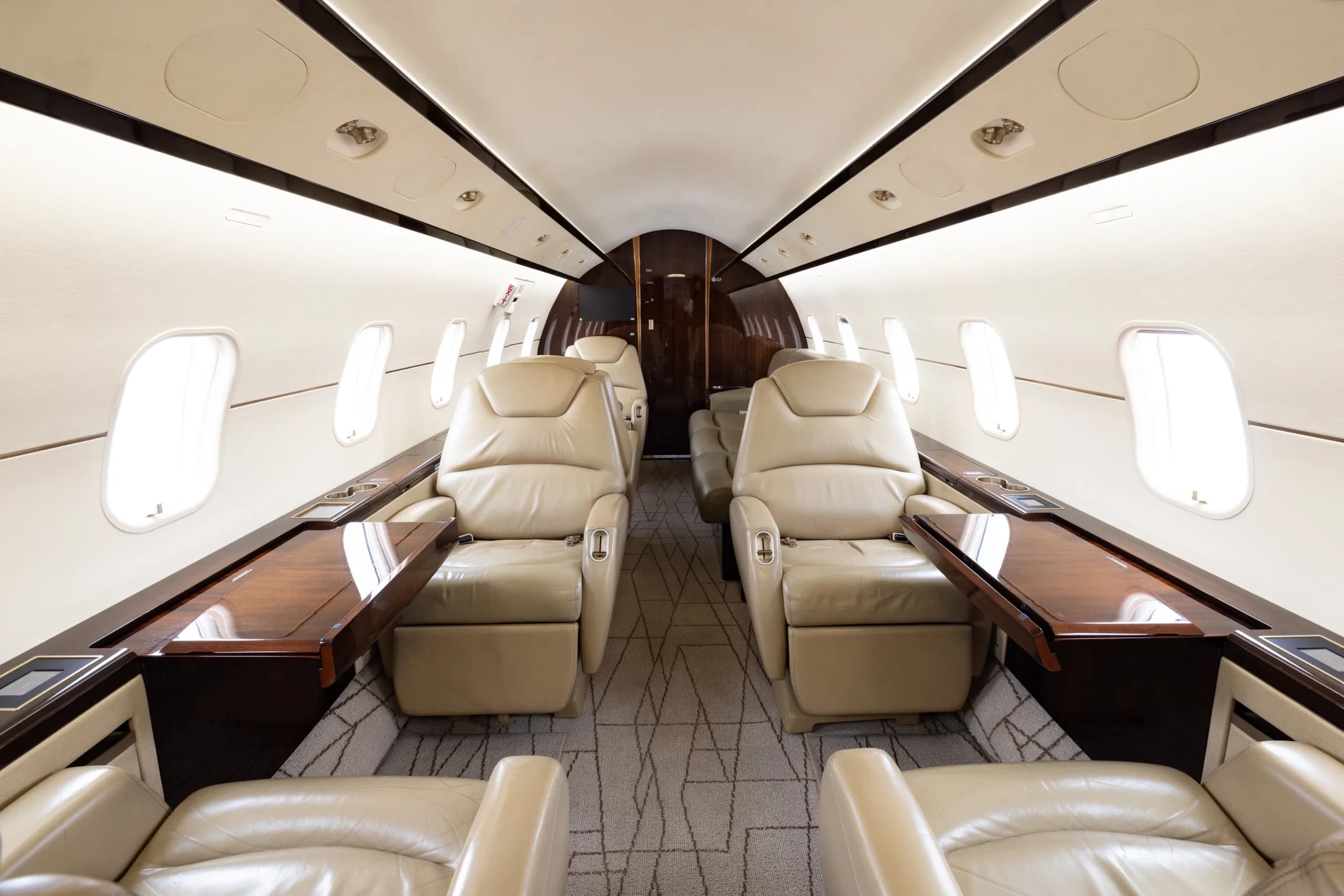 The luxurious interior of a business jet cabin with light beige leather seats and dark wood tables.