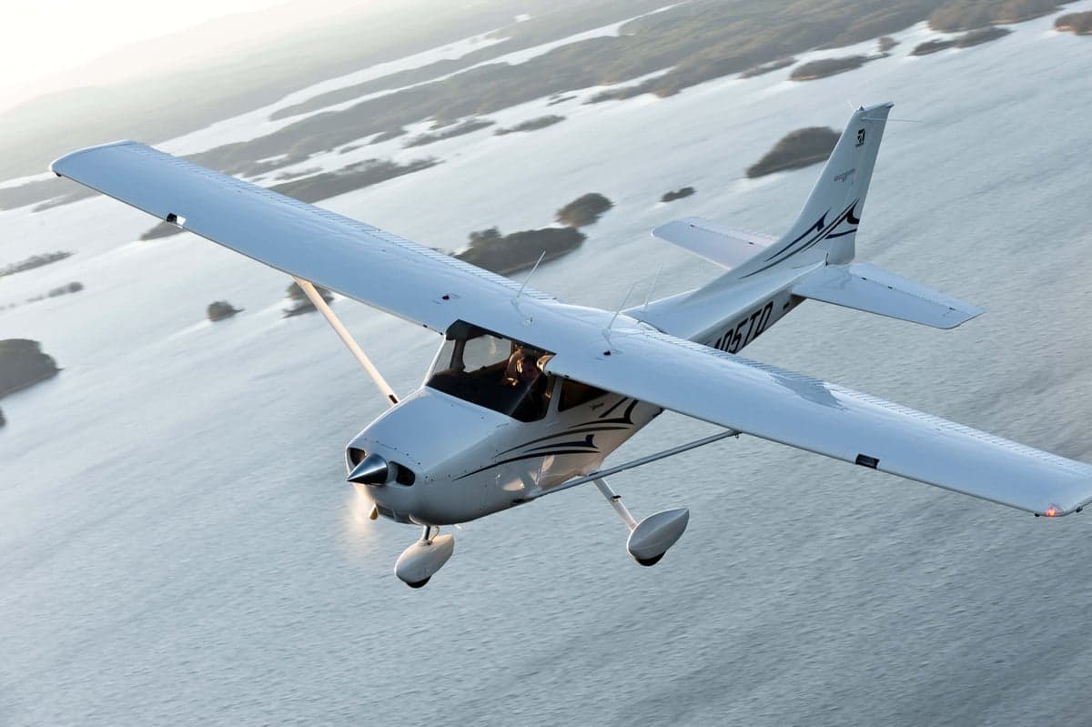 A white Cessna 182 aircraft with black stripes flying over a body of water with scattered islands.