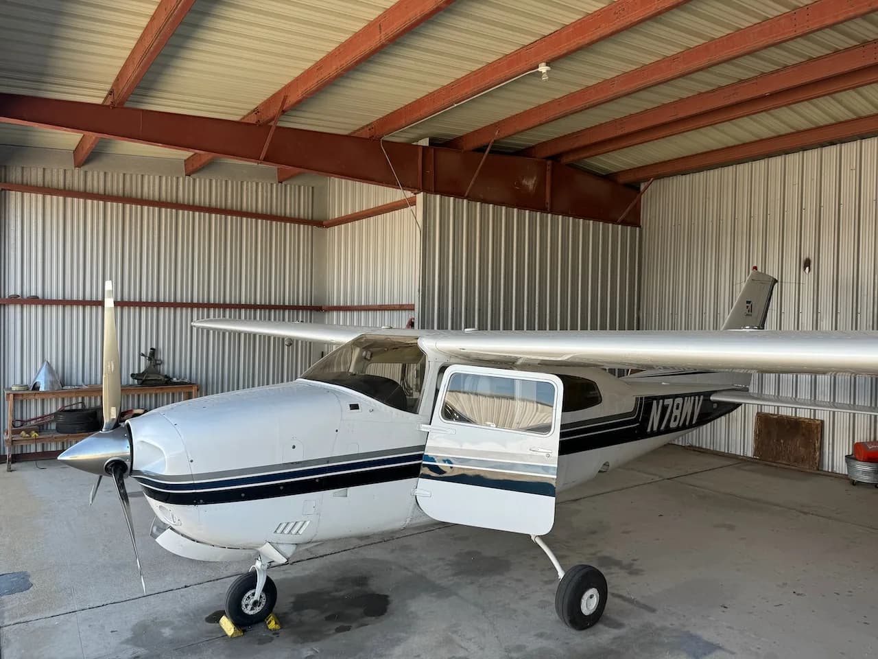 A white Cessna aircraft with blue and black stripes, registration N78WV, with its door open, parked in a hangar.