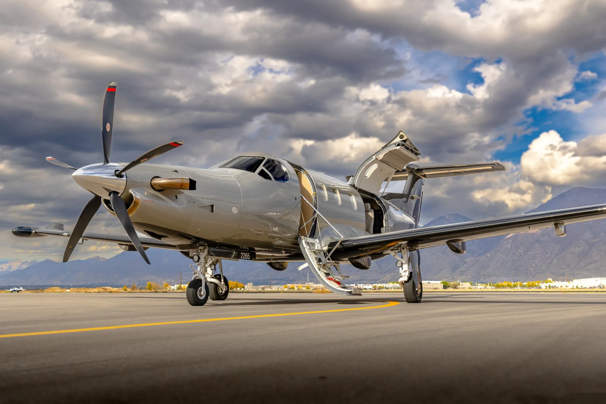 A silver Pilatus PC-12 turboprop aircraft with its main and cargo doors open, parked on an tarmac with mountains in the background.