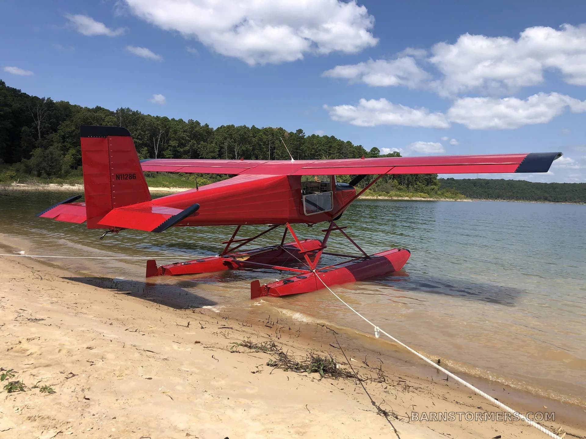 A red floatplane with registration N11286 is tied to a sandy beach, partially submerged in a lake under a blue sky.
