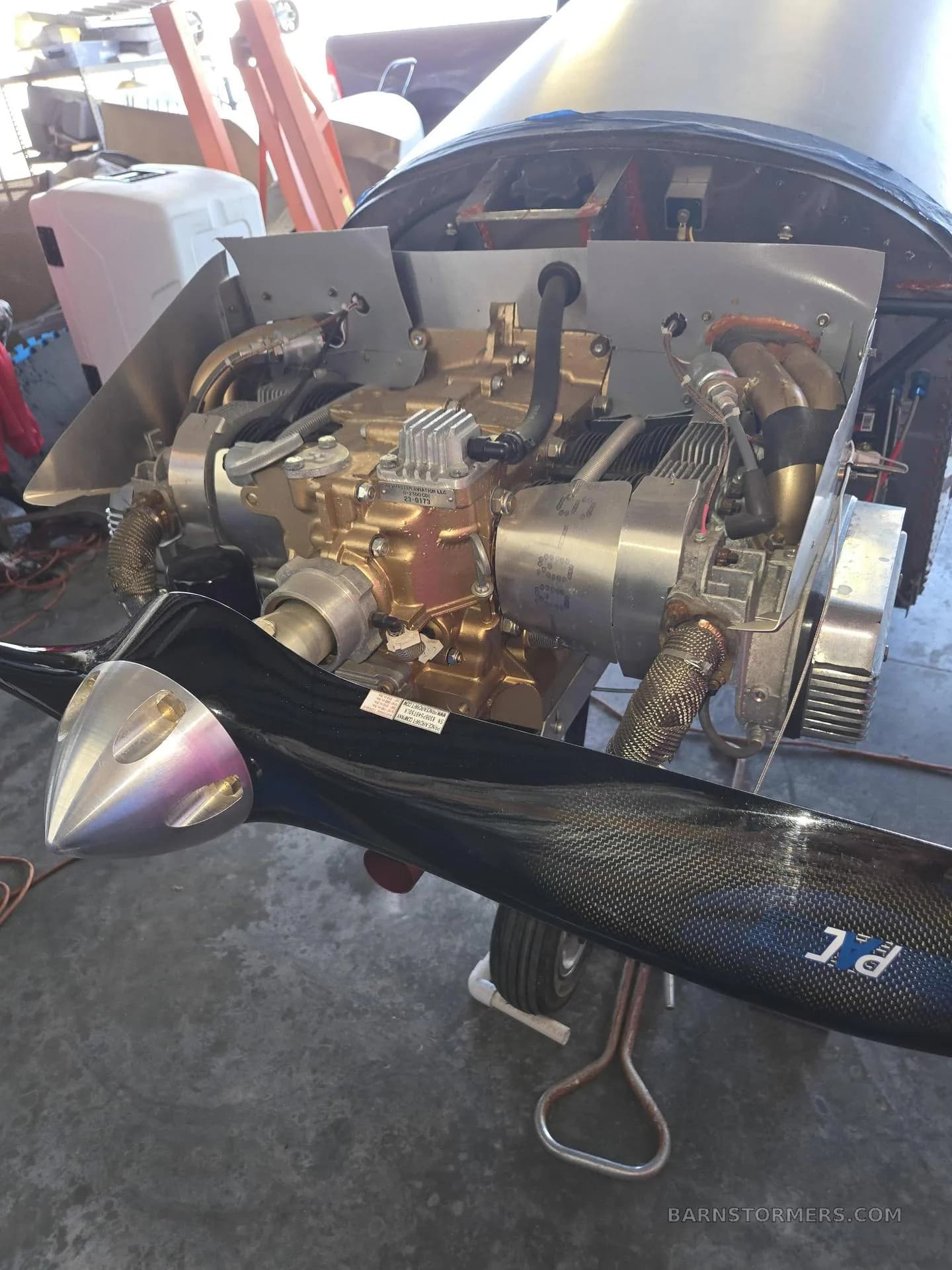 A front view of an aircraft engine with a black propeller, in a workshop setting.