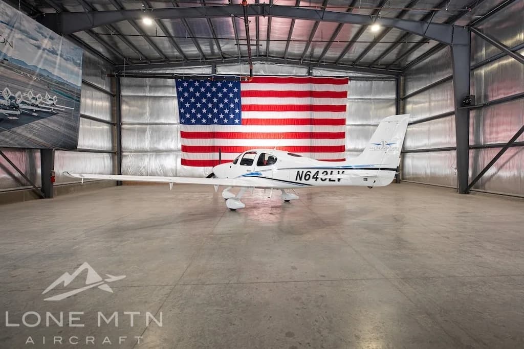 A white Cirrus SR22 aircraft with blue and black stripes, tail number N643LV, parked in a hangar with an American flag in the background.