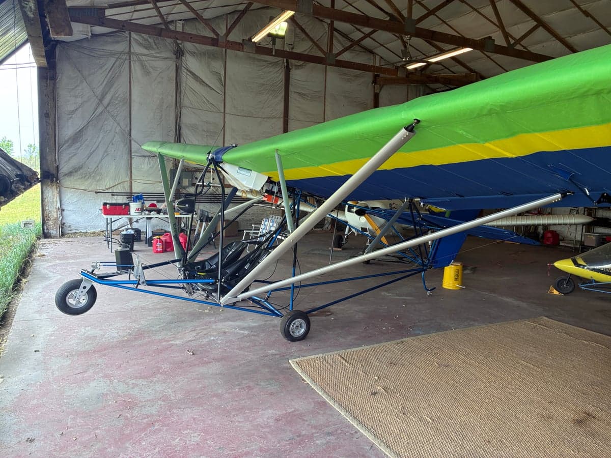 Side view of a blue, green, and yellow ultralight aircraft with an exposed frame, parked inside a hangar.