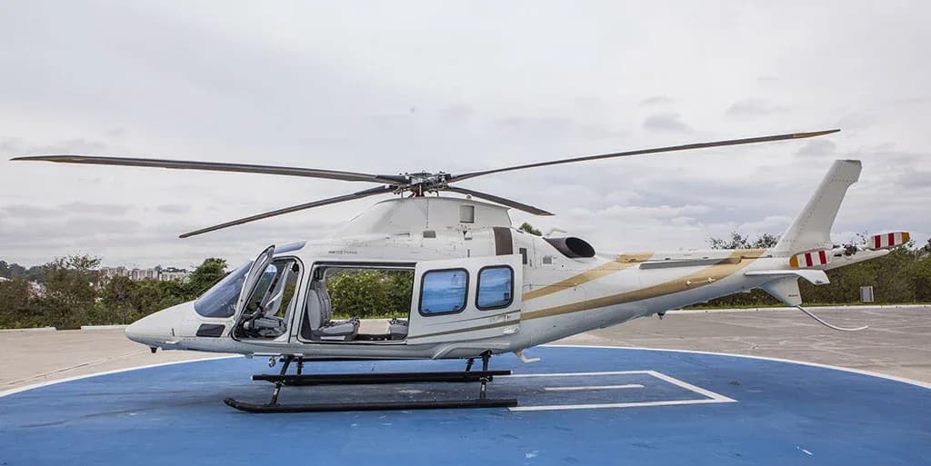 A white helicopter with gold stripes is parked on a blue helipad with its cabin doors open, viewed from the side.