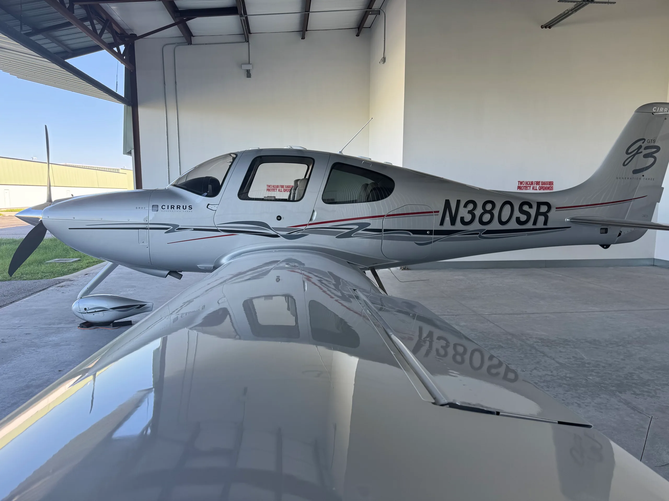A silver Cirrus SR22 Generation Three aircraft with N380SR registration is parked in a hangar, viewed from the front-left.
