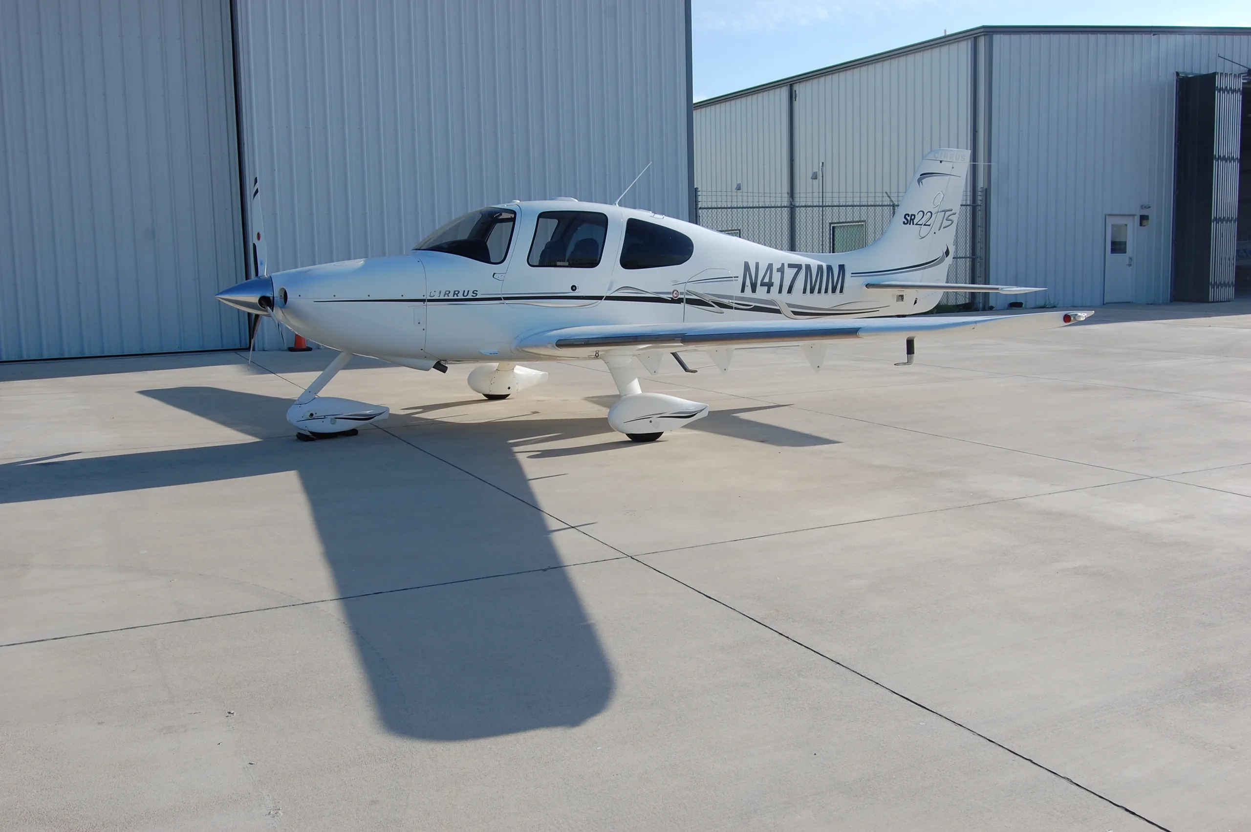 A white Cirrus SR22 aircraft, registration N417MM, parked on an airfield in front of hangars.