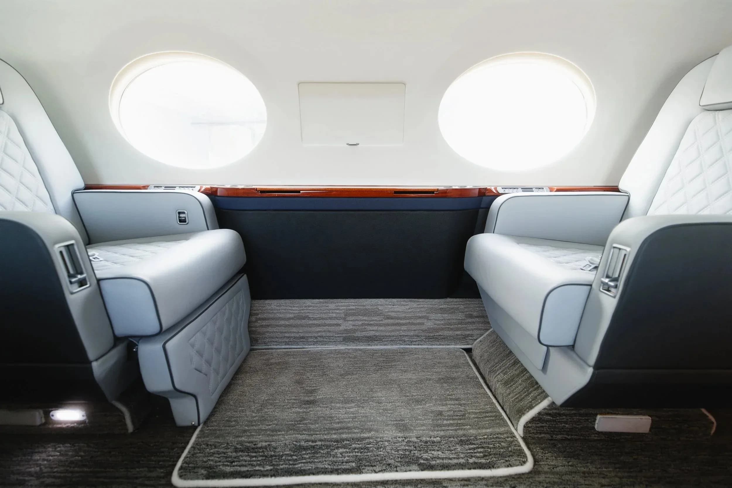 Two light gray quilted leather seats facing each other in the private jet's cabin, with large circular windows.