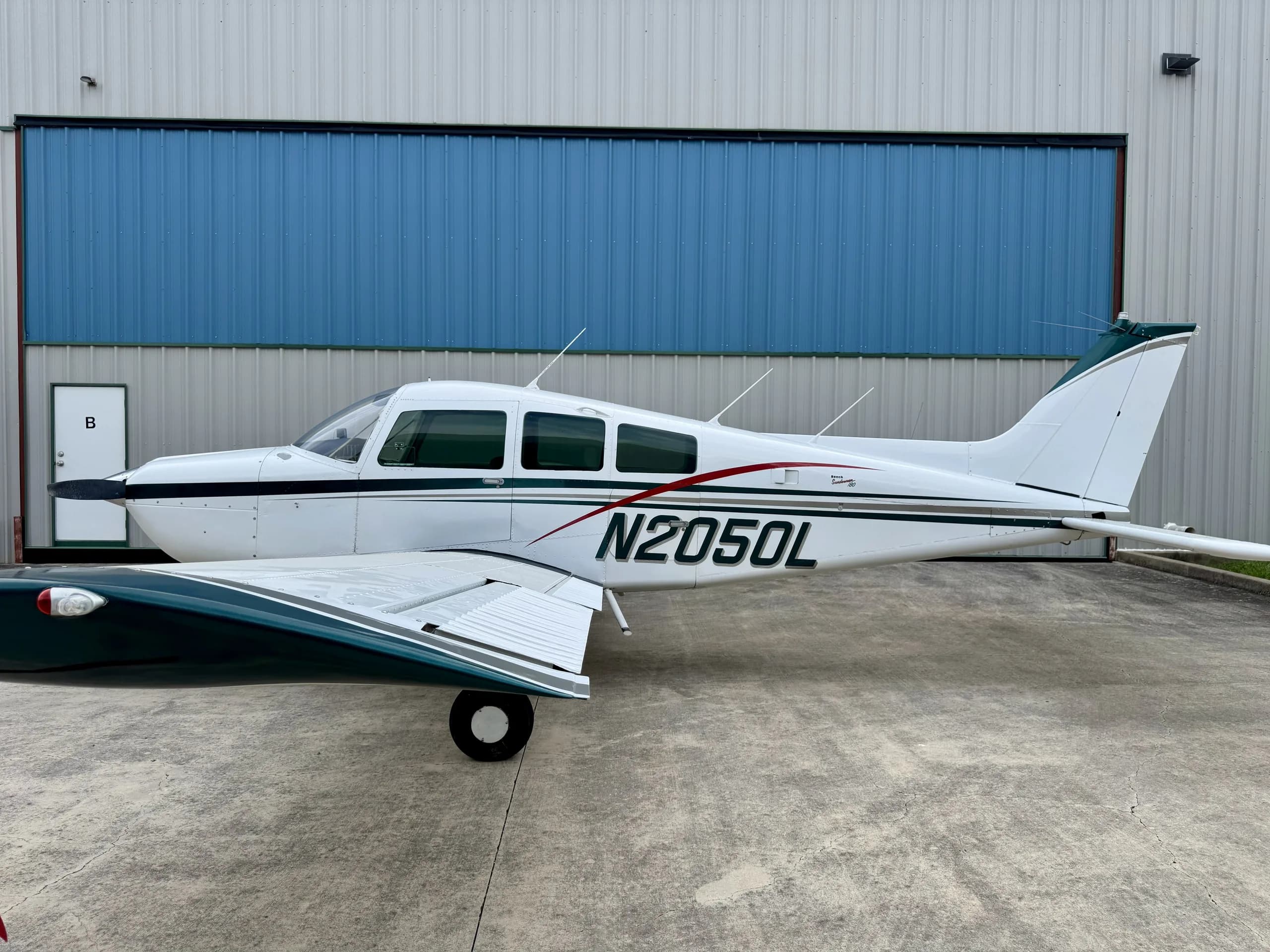 Side view of a white and green Beechcraft Sundowner N2050L parked in front of a hangar.