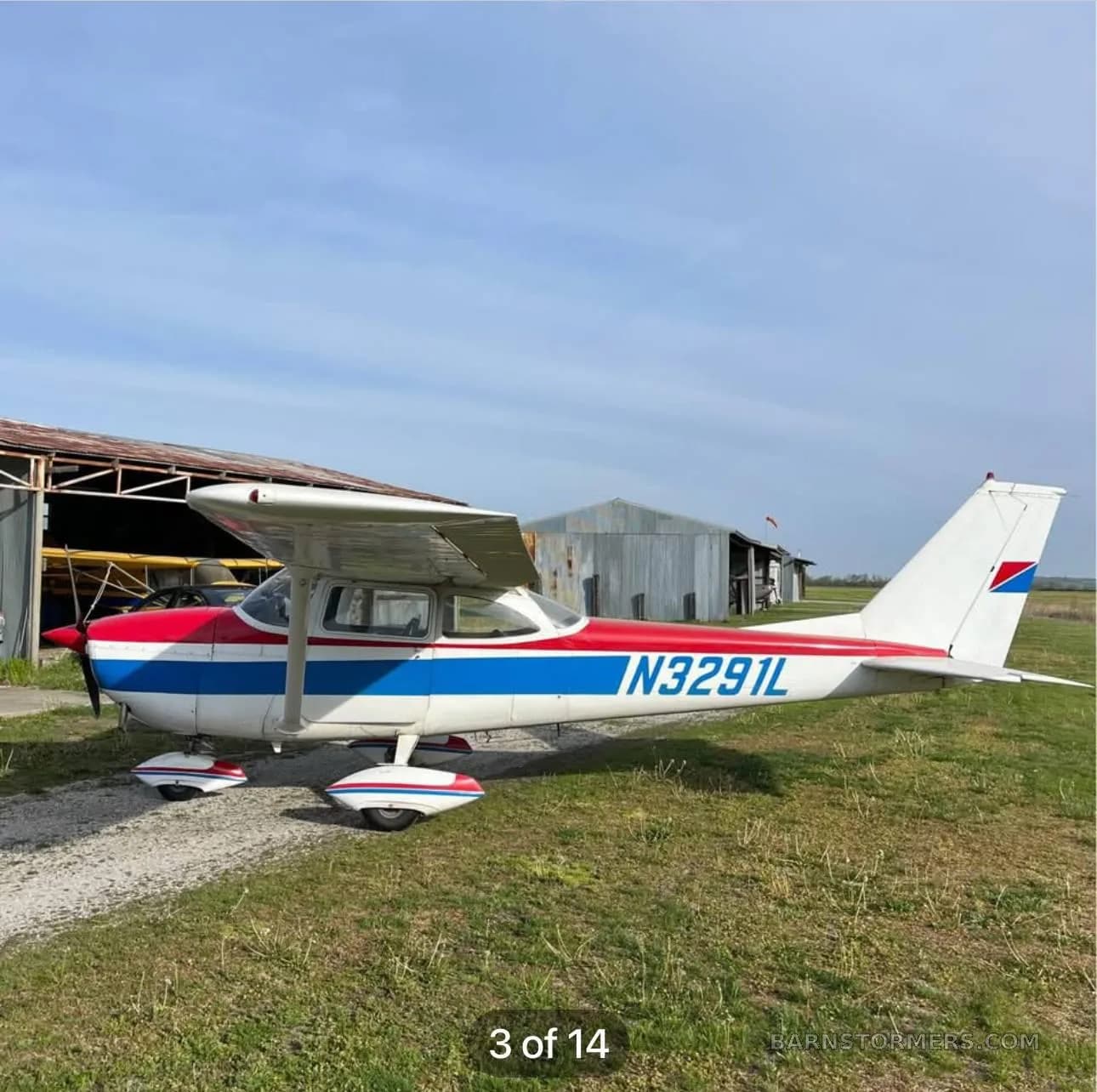 A side view of a white, red, and blue Cessna-style aircraft with registration N3291L parked on a gravel path.