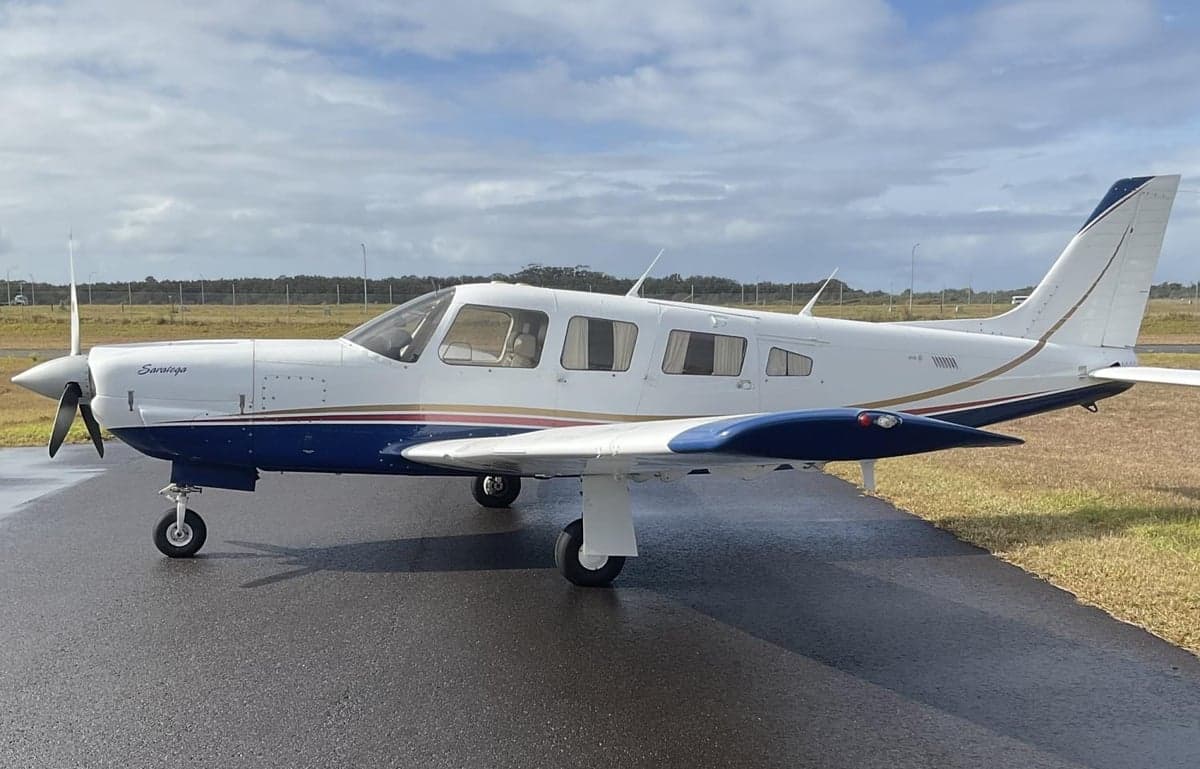 A white and blue Piper Saratoga aircraft with brown and gold stripes is parked on a wet tarmac under a cloudy sky.