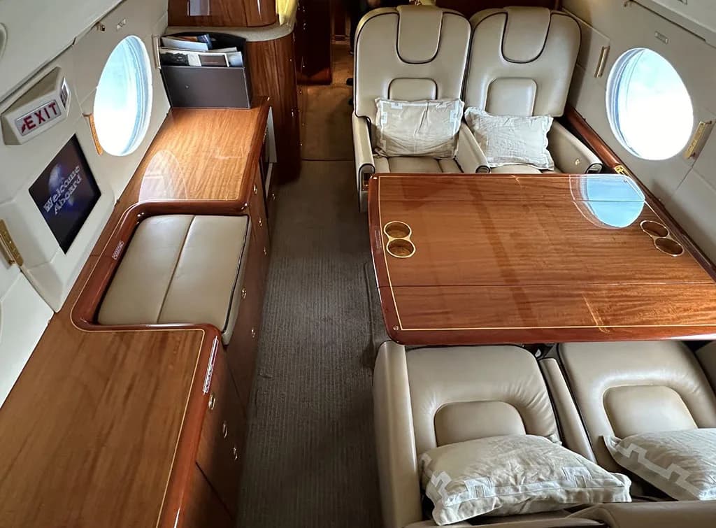 A private jet cabin interior showing a galley counter with a screen and a dining area with beige seats and a large wooden table.