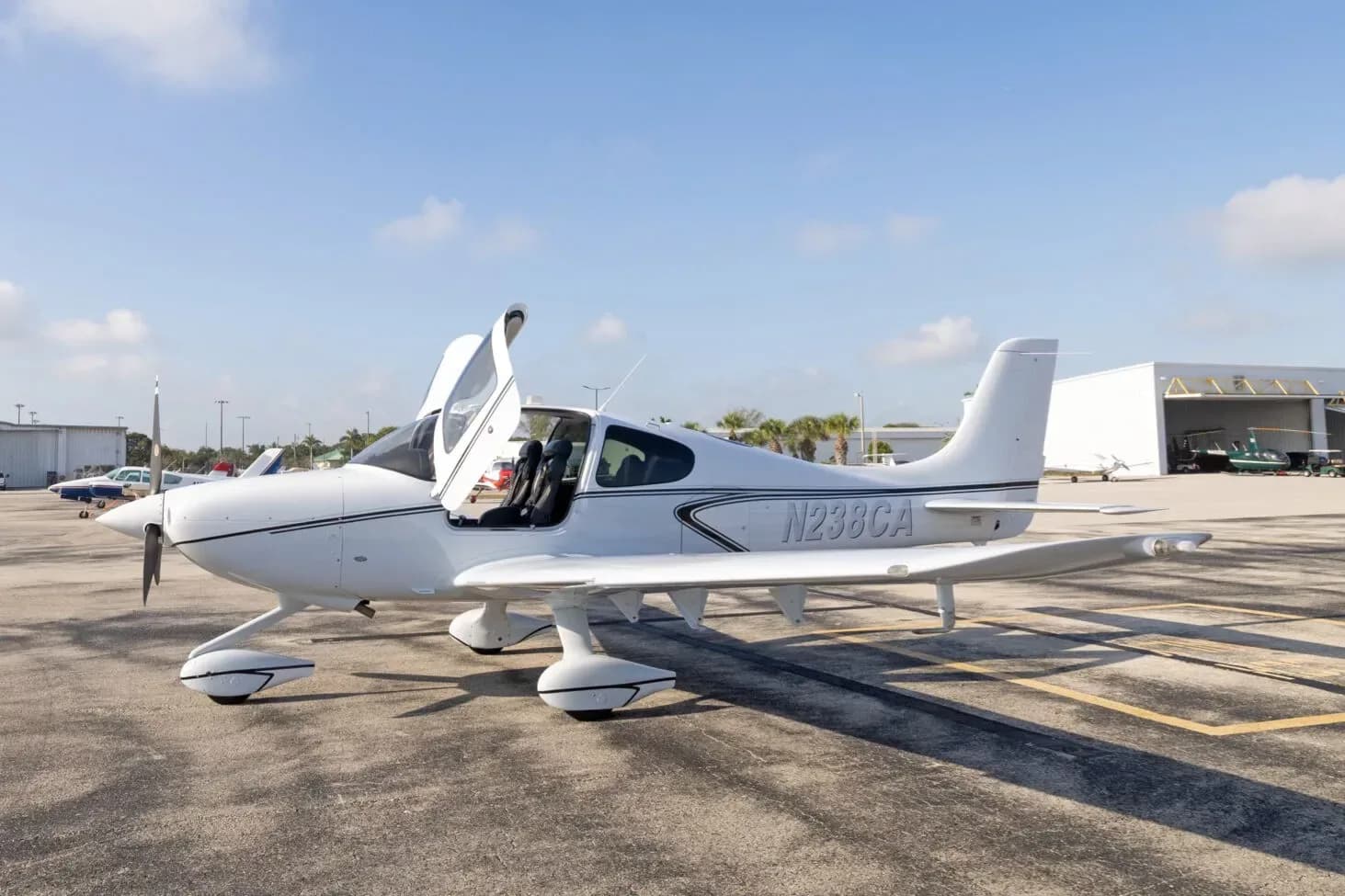 A white Cirrus SR22 aircraft with its gull-wing doors open is parked on an airport tarmac near a hangar.