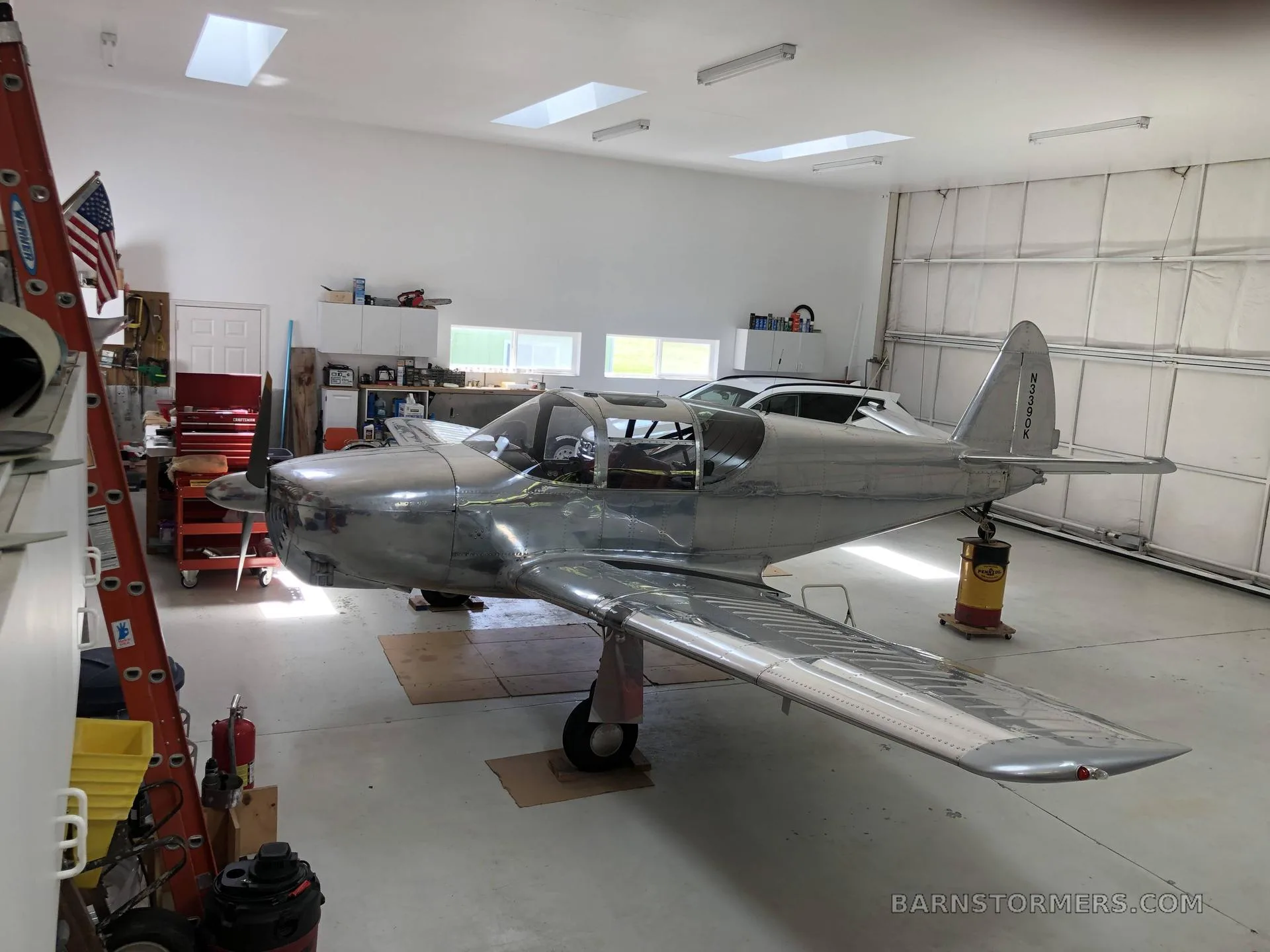 A shiny silver single-engine aircraft is parked inside a bright hangar, viewed from the front-left.
