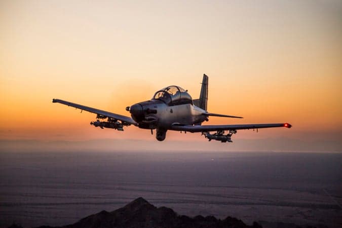 An AT-6 light attack aircraft flying at sunset over a desert landscape.